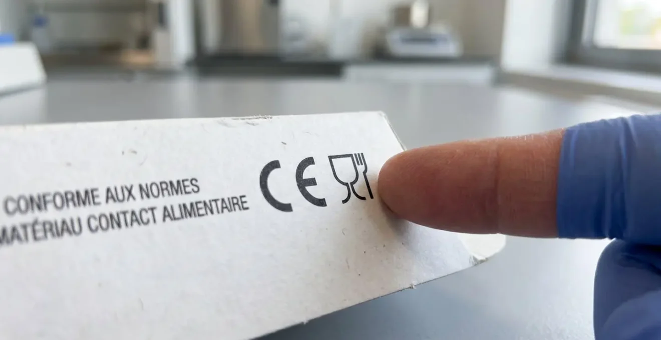 La main d'un professionnel examine le marquage de conformité imprimé au dos d'un plateau en carton blanc dans un laboratoire de pâtisserie